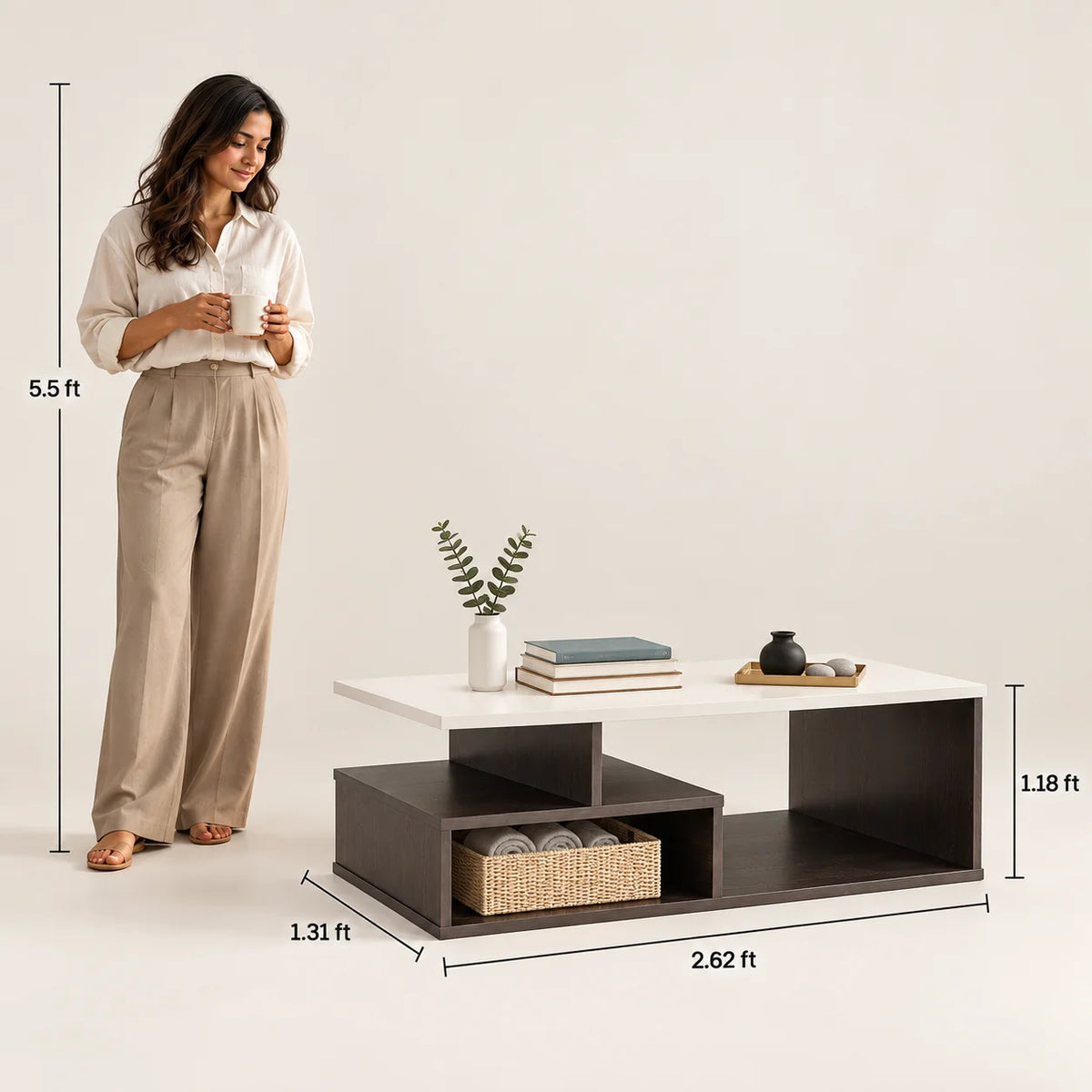 A woman stands beside a modern low coffee table with overlaid dimension lines showing its height, width, and depth. The table has a white top surface and dark brown open shelving underneath, with decorative books, a small plant, and a tray on top.