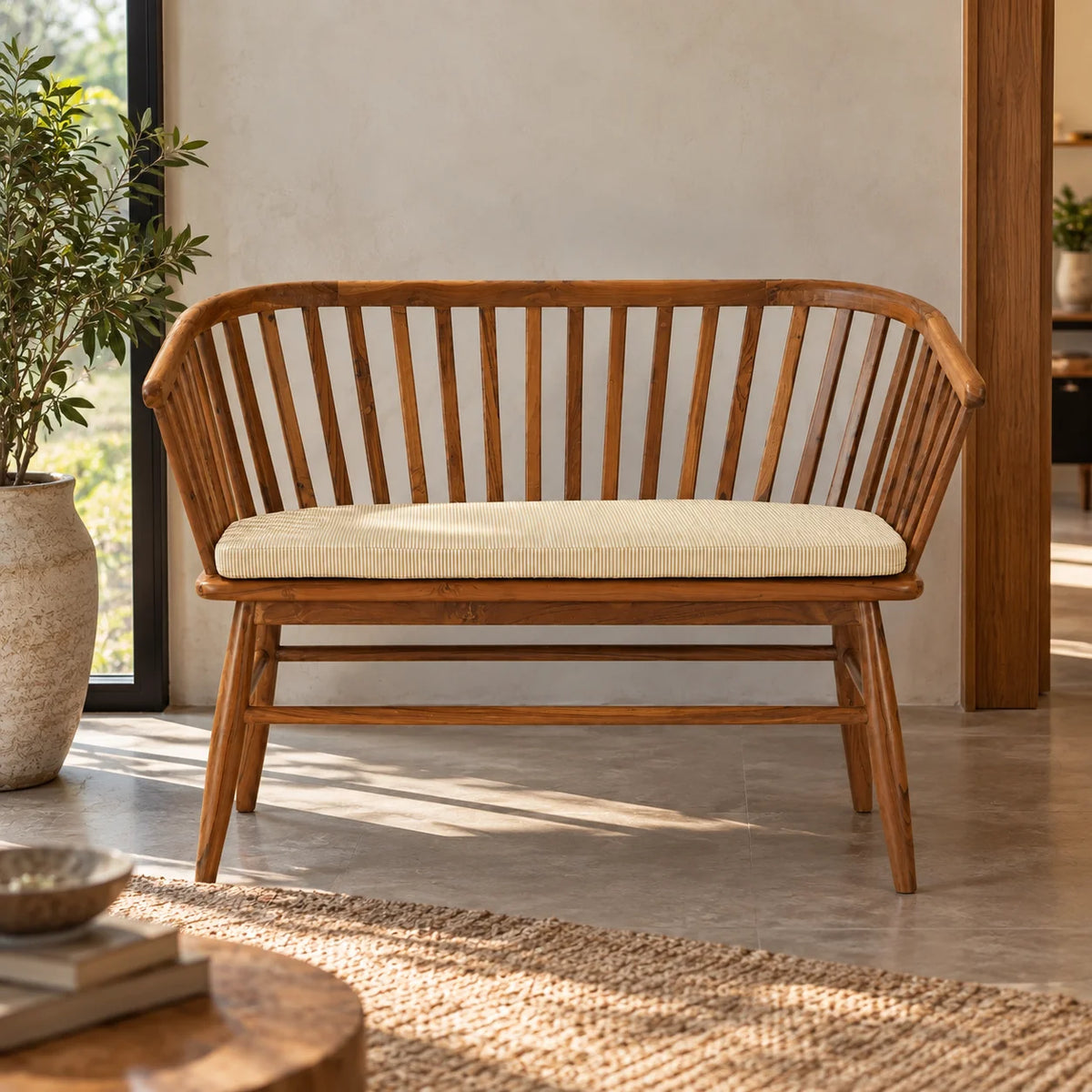 A wooden bench with a curved slatted backrest and a cream-colored cushion, placed in a bright, modern room with a concrete floor, a potted plant, and a woven rug.