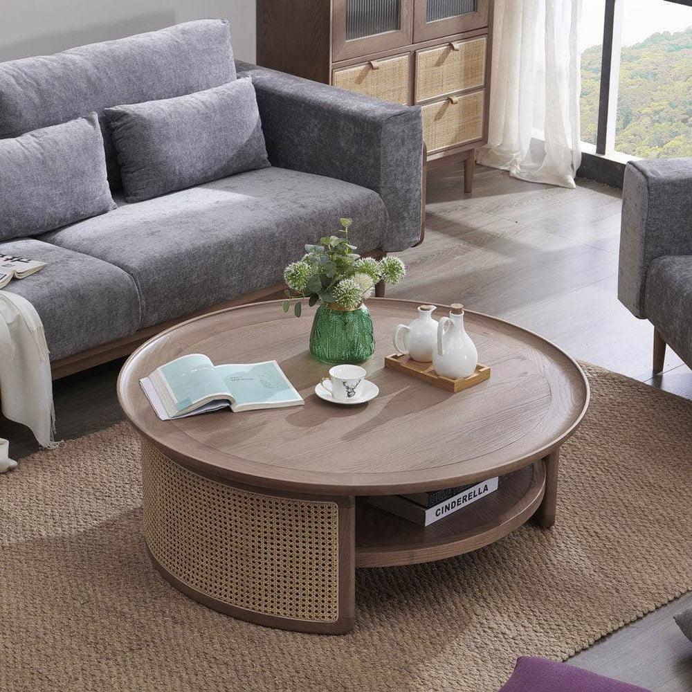 A round Pluto coffee table made of mango wood with a rattan base, featuring a natural wood top and brown color, placed in a living room setting with a grey sofa and decorative items on top.